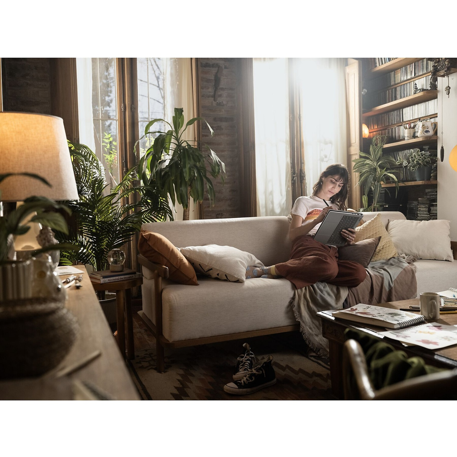Een vrouw zit op een lichte bank in een zonnige woonkamer met planten en tekent op een tablet.