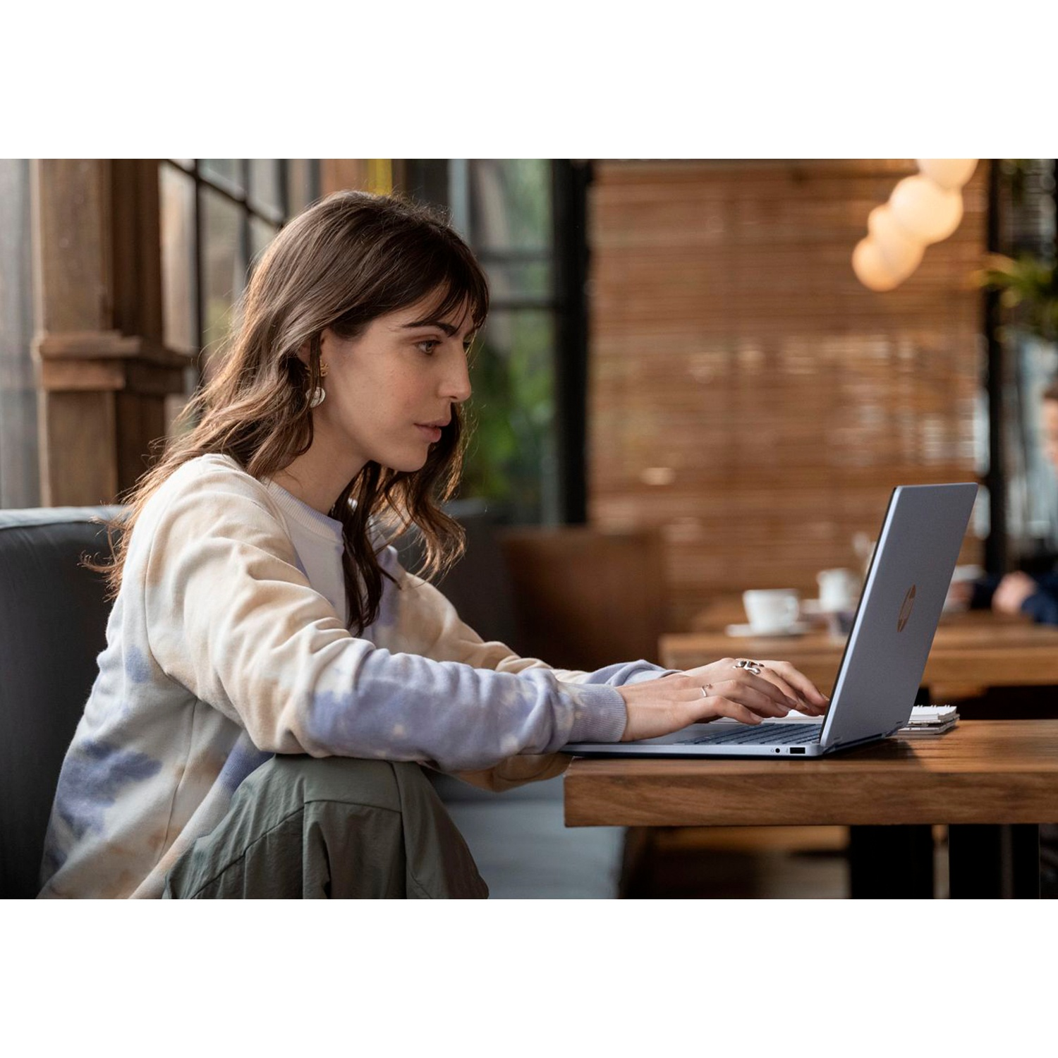 Een vrouw in een truien met patroon zit aan een houten tafel en typt geconcentreerd op een laptop.