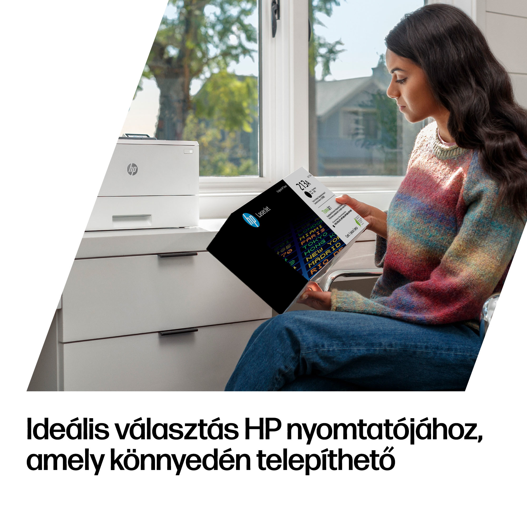 Egy pulóveres nő HP toner dobozt tart az ablak mellett, mellette egy nyomtató áll a szekrényen.