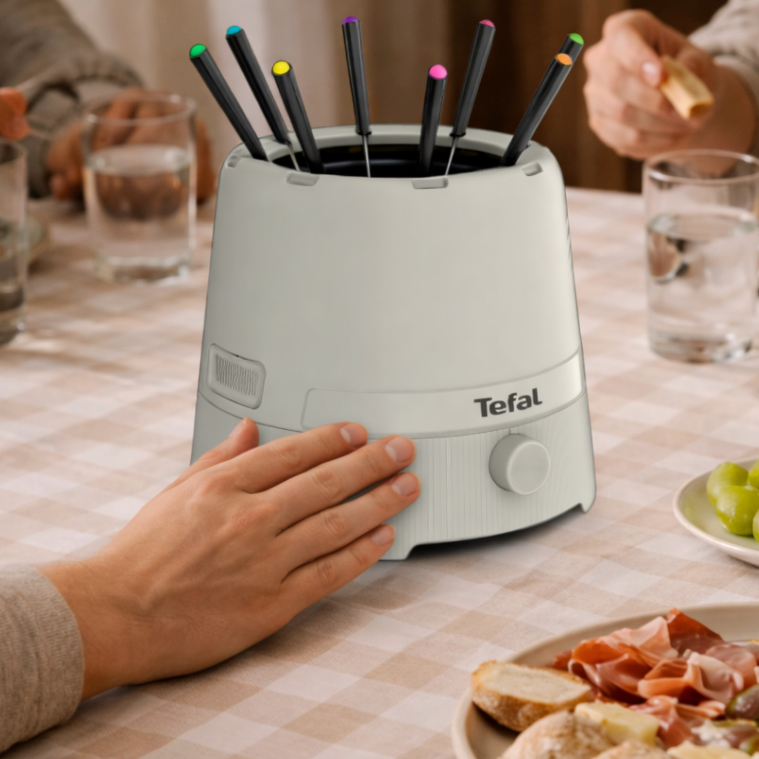 Een hand raakt een witte Tefal fonduepan met gekleurde vorkjes aan.