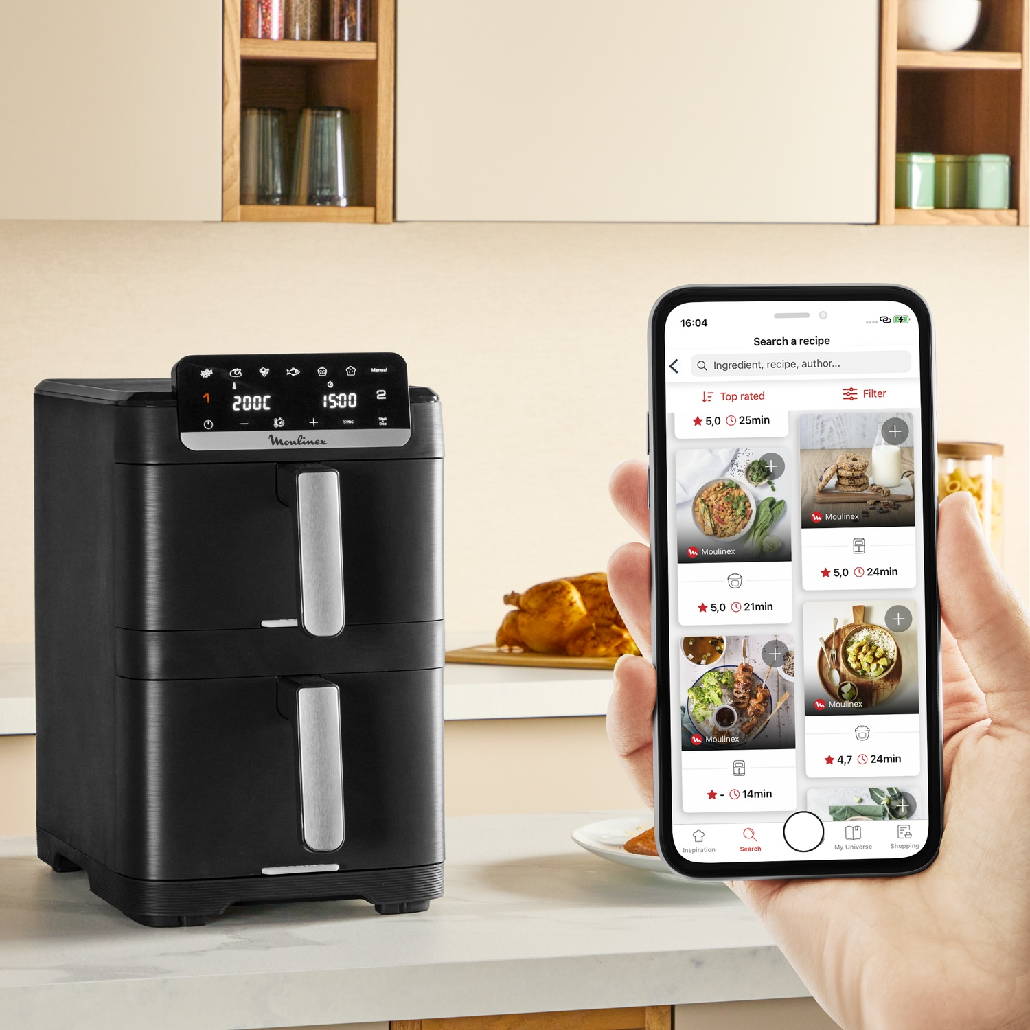 Friteuse sans huile noire sur un plan de travail avec une main tenant un smartphone affichant des recettes.