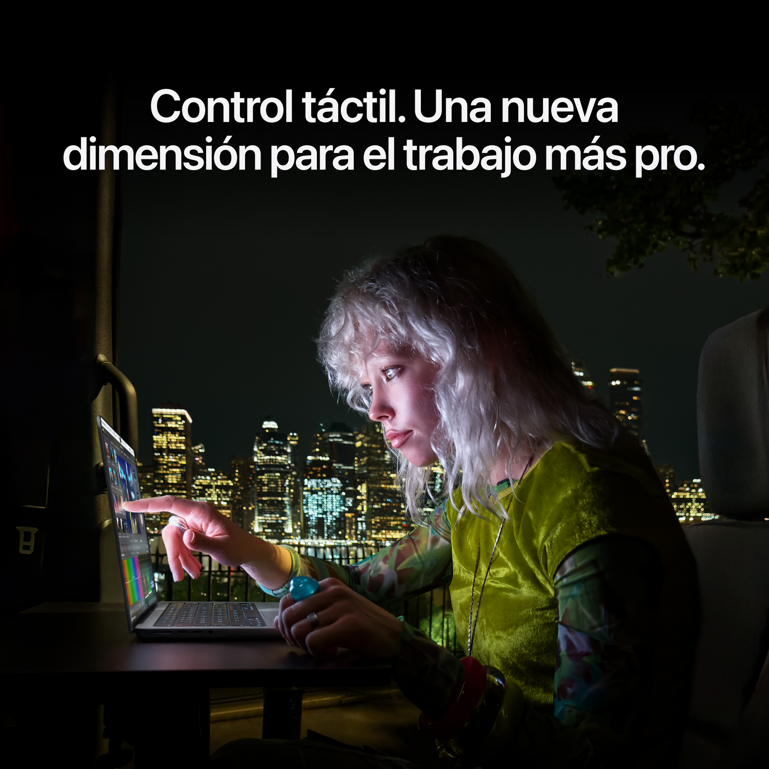Una mujer rubia usa la pantalla táctil de una laptop de noche, con un horizonte de ciudad iluminado de fondo.