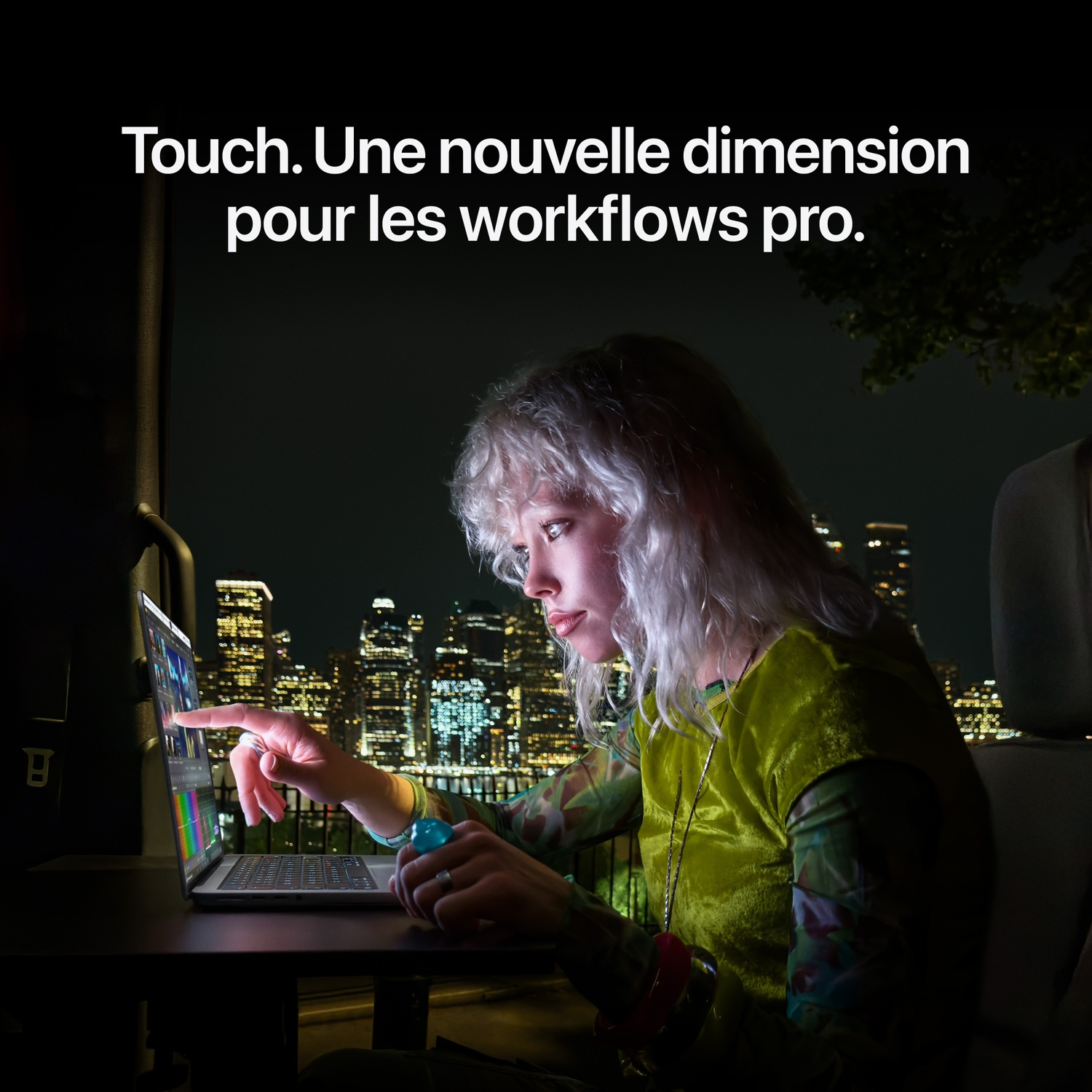 Une femme aux cheveux clairs utilise un ordinateur portable à écran tactile la nuit devant une ville illuminée.