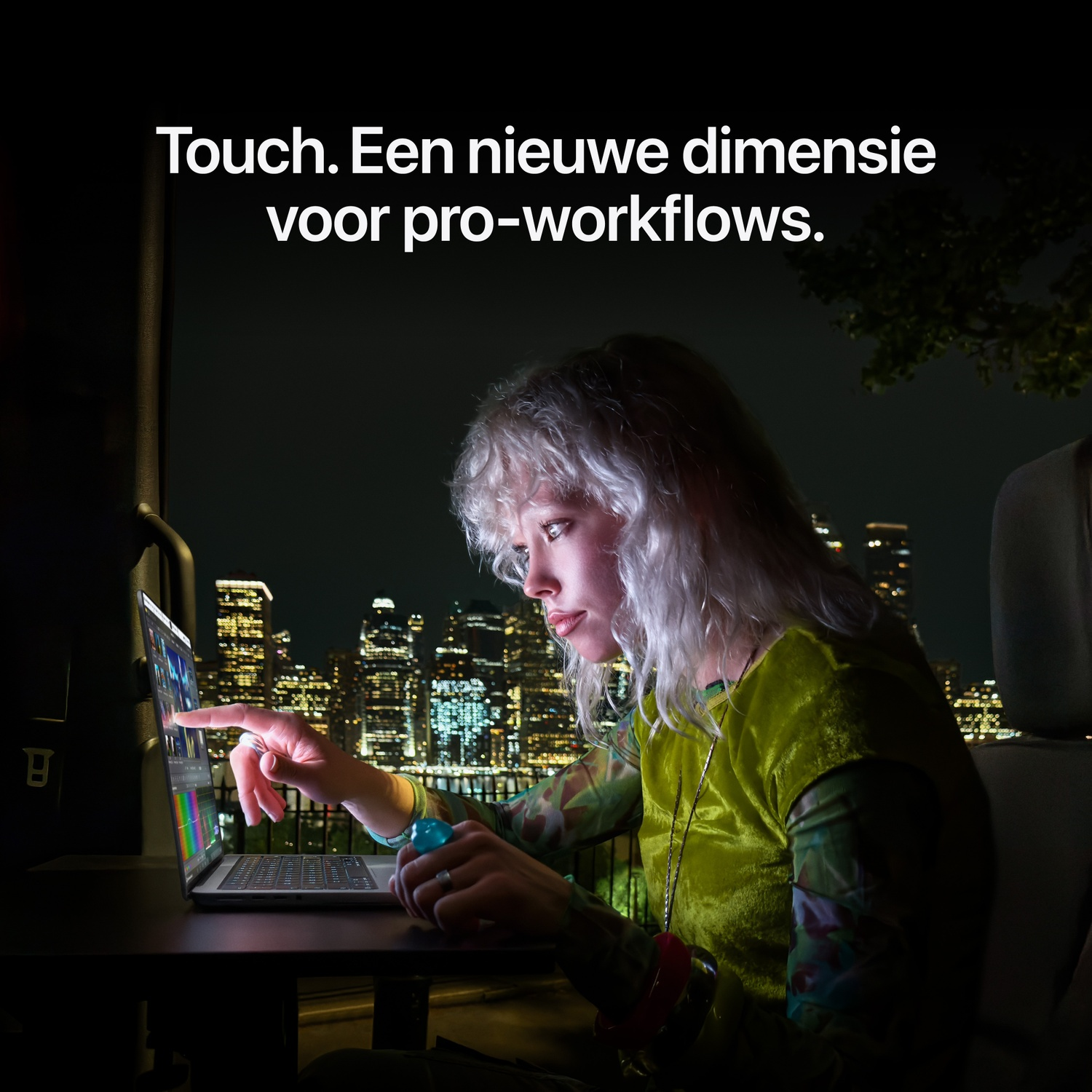 Een blonde vrouw gebruikt 's nachts in een auto het touchscreen van een laptop met stadsgezicht op de achtergrond.