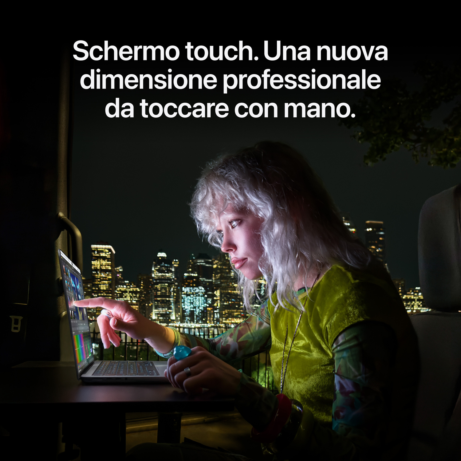 Donna usa il touchscreen di un laptop in auto di notte, con uno skyline cittadino illuminato sullo sfondo.