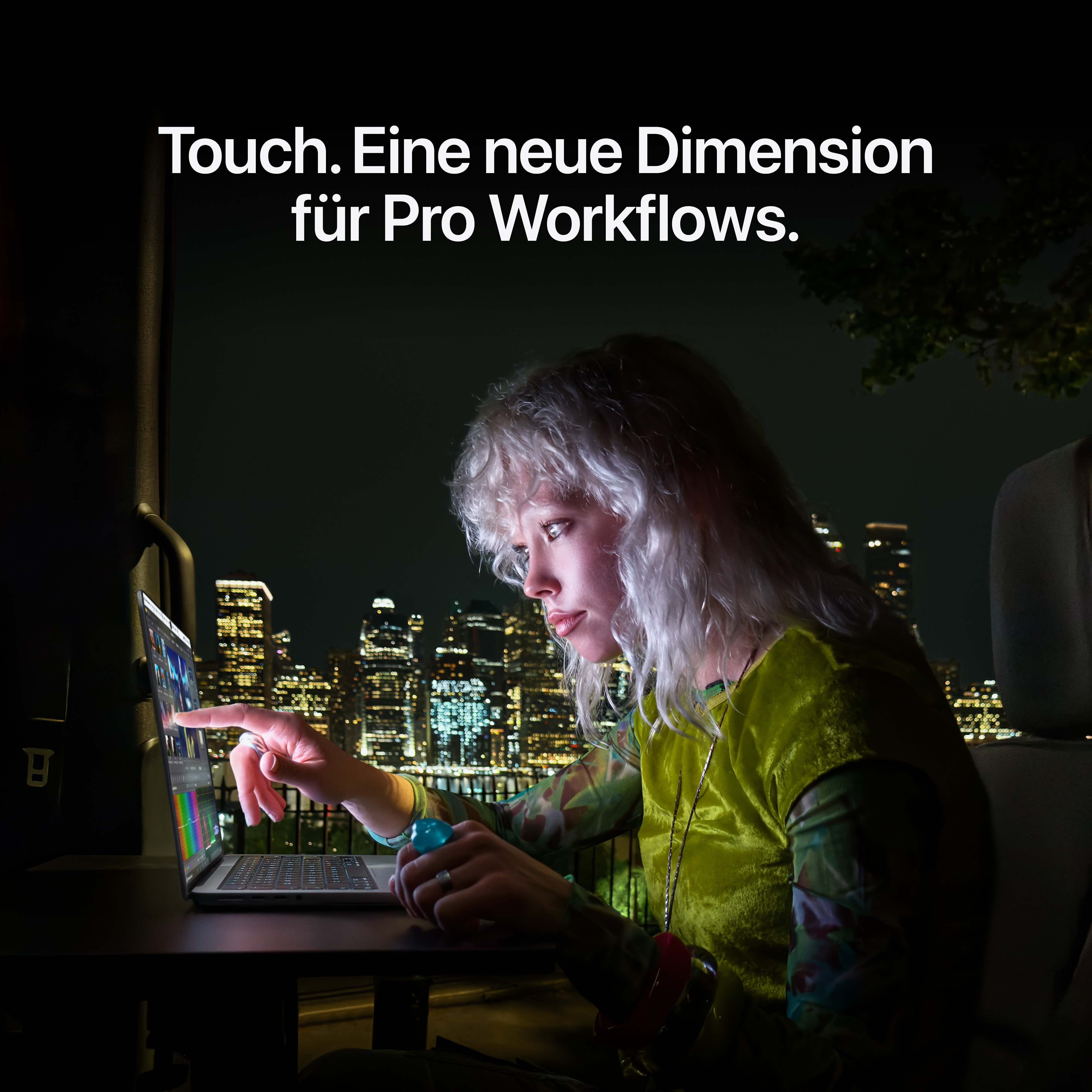 Eine Frau mit heller Haut und hellem Haar tippt nachts auf den Touchscreen eines Laptops, im Hintergrund die Stadt.
