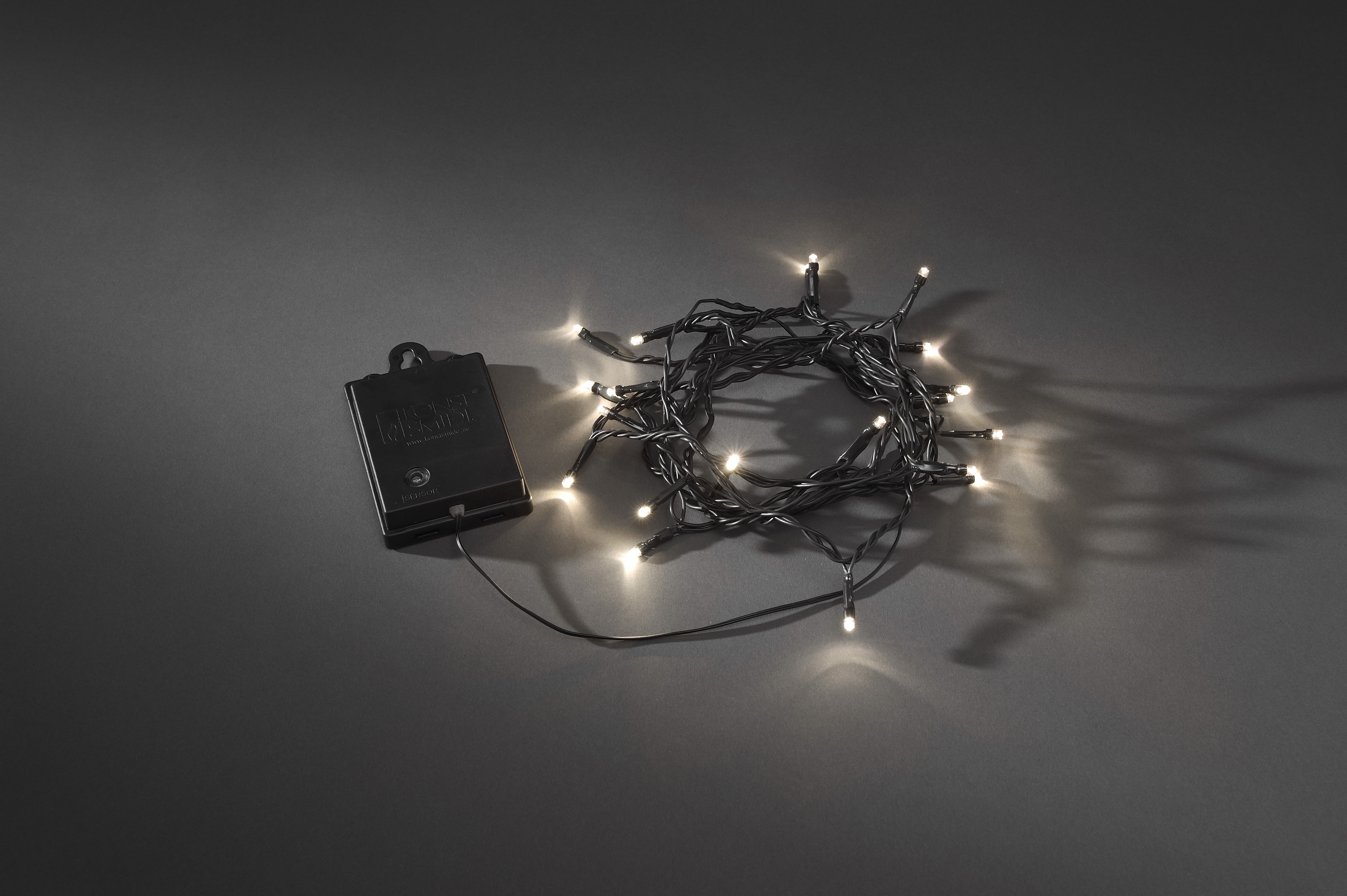 Eine Lichterkette mit kleinen weißen Lichtern und einem schwarzen Akku-Pack auf dunklem, grauem Hintergrund.