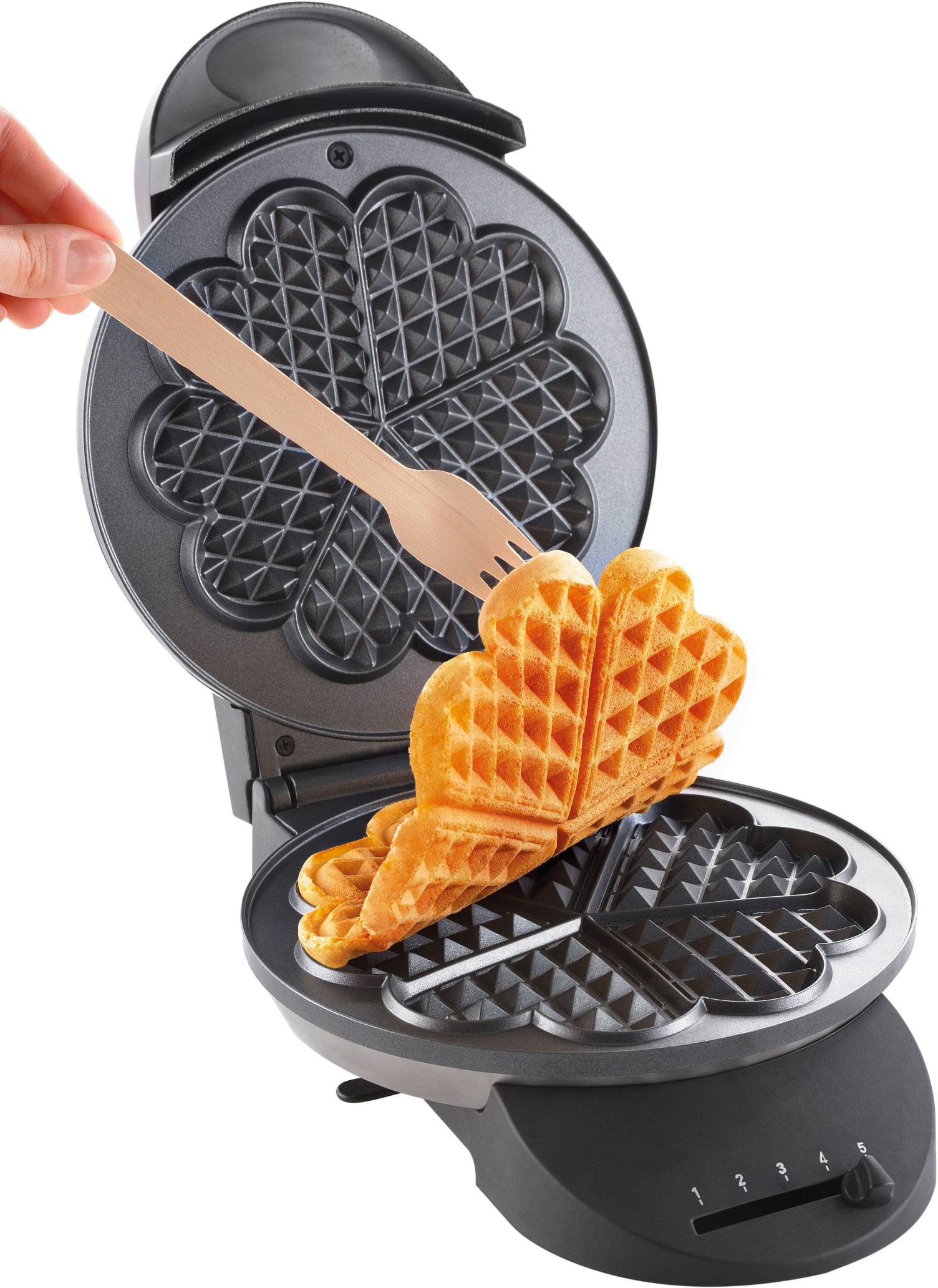 Eine Person, die mit einer Holzgabel ein herzförmiges Waffel aus dem Waffeleisen nimmt.
