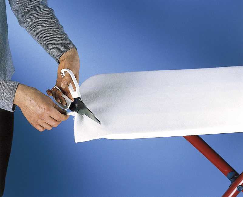 Personne coupant du tissu blanc avec des ciseaux sur une planche à repasser, fond bleu.