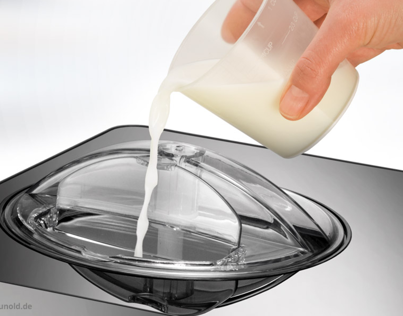 Eine Hand gießt Milch aus einem Messbecher in einen durchsichtigen Kunststoffbehälter.