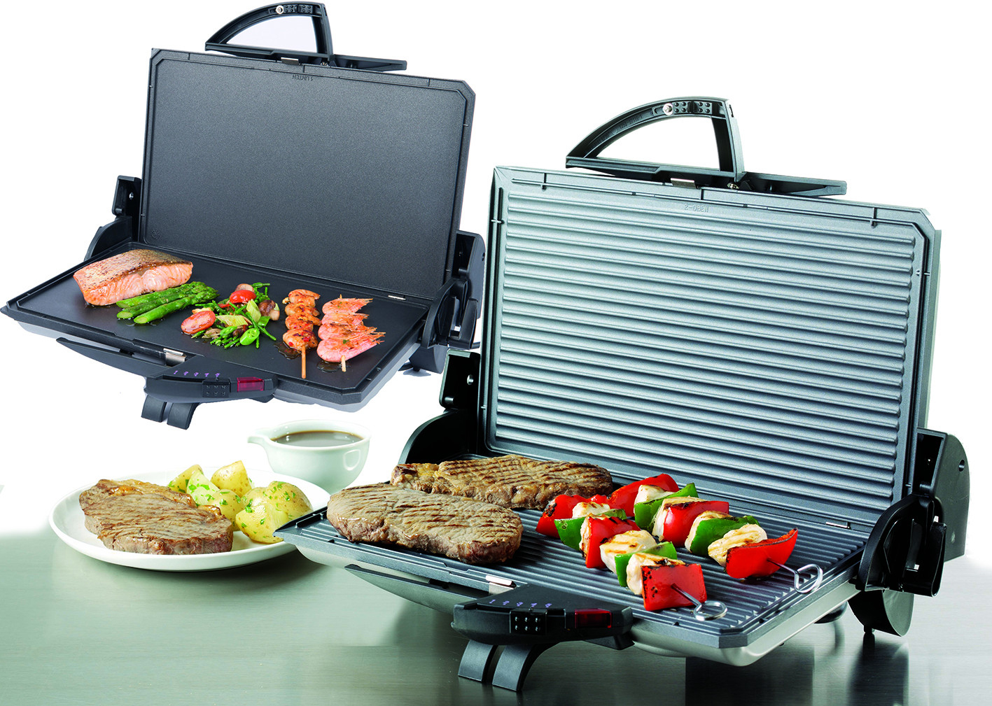 Zwei offene Elektrogrills. Einer mit gekochtem Essen, darunter Fleisch, Gemüse und der andere mit Steaks.