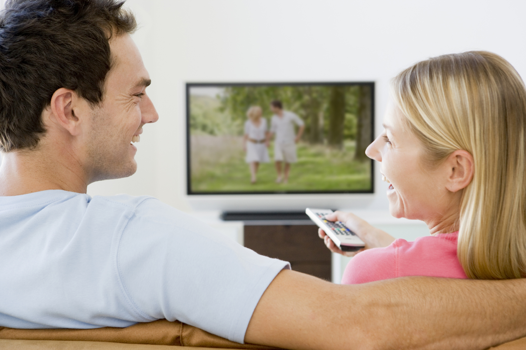 Couple watching TV. Man and woman are smiling. The TV shows a couple walking in a green field.