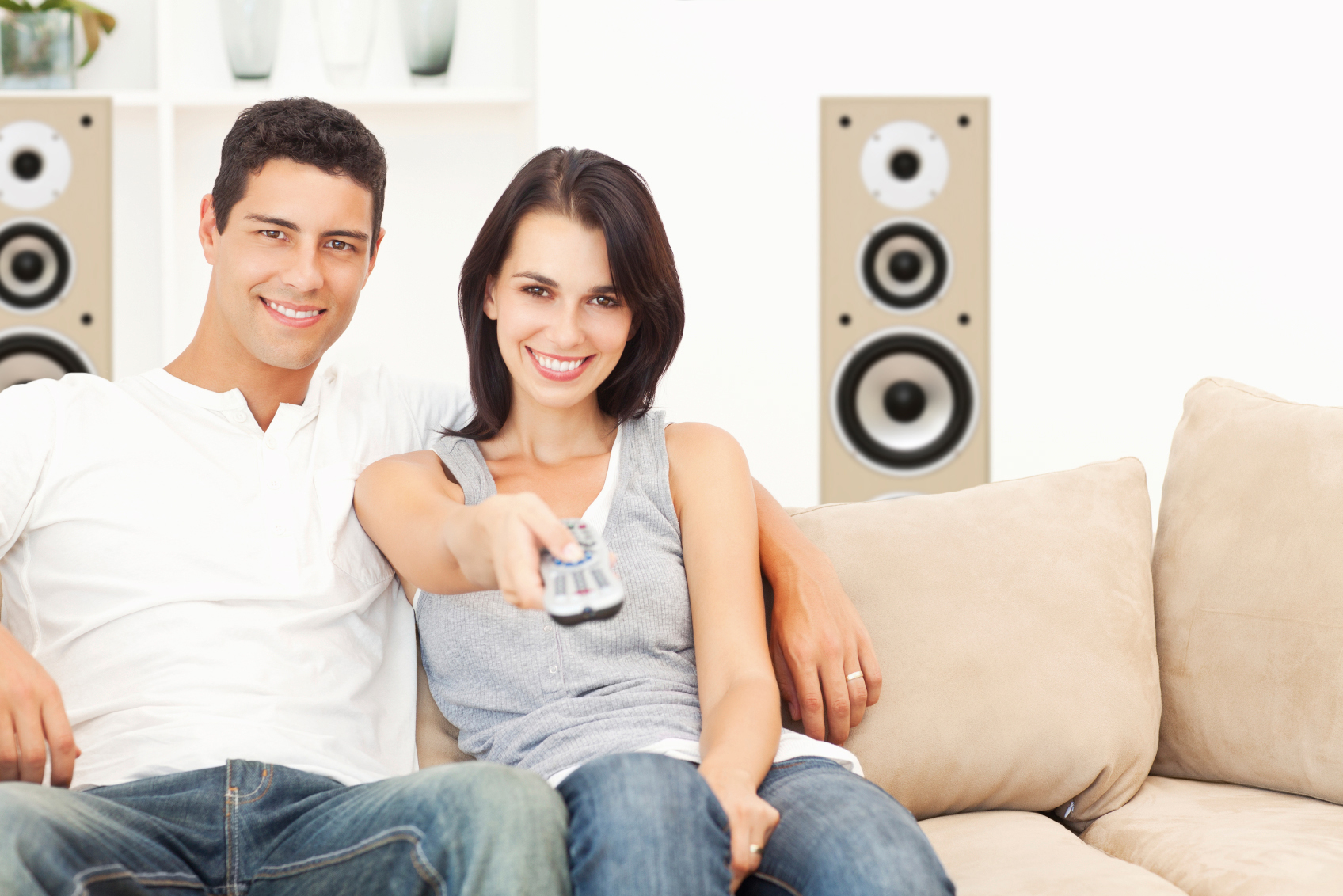 A couple smiles on a couch. The woman points a remote. Speakers are in the background.