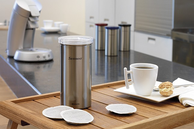 Een koffiezetapparaat, een container, koffiepads en een kopje op een houten dienblad.