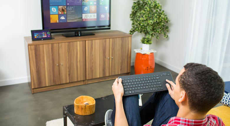 Bir adam bir klavye kullanıyor ve bir TV ekranına bakıyor, bir dolapta bir tablet var. Parlak oda.
