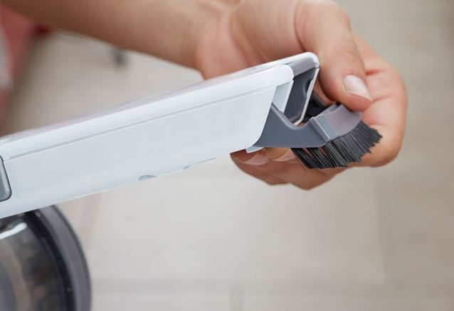 A hand holds a brush attachment for a white and gray vacuum cleaner.