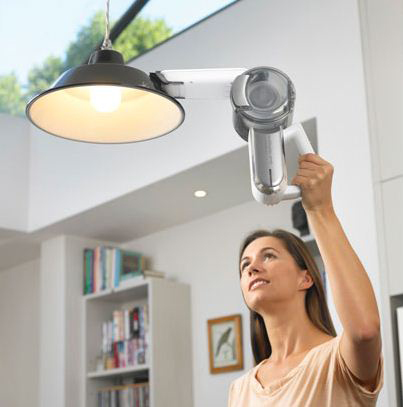 A woman vacuuming a lampshade with a handheld vacuum cleaner, in a well-lit, modern interior.