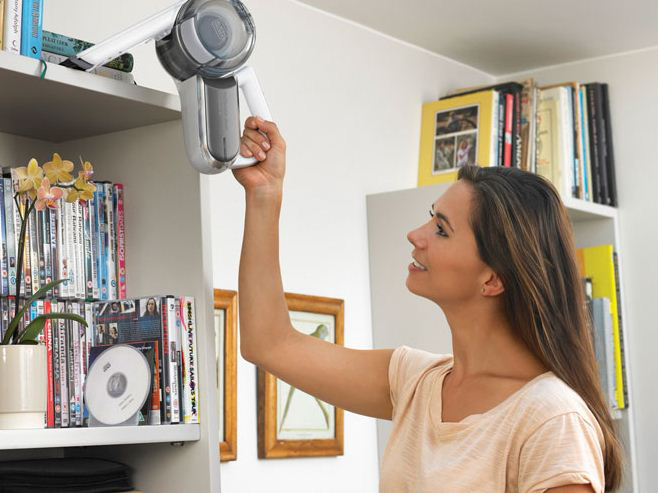 A woman using a cordless vacuum cleaner to clean a bookshelf.