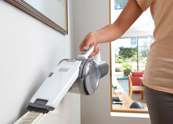 A person is using a cordless vacuum cleaner on a white wall with a radiator.