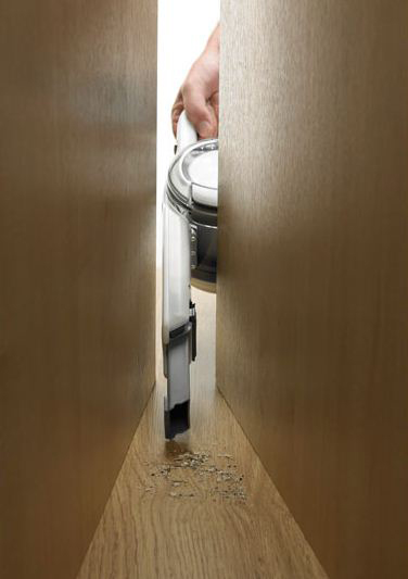 A vacuum cleaner being used in a narrow space between two pieces of wood. The floor is visible.