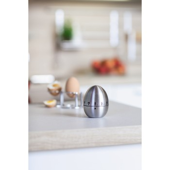 A stainless steel egg timer on a counter. Two eggs in holders and a broken egg are in the background.