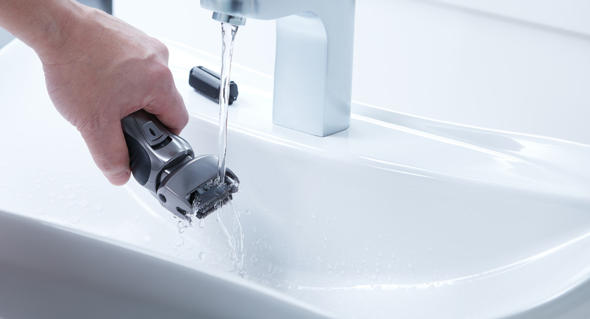 Die Hand eines Mannes, die einen schwarz-grauen Elektrorasierer unter einem Wasserhahn wäscht.