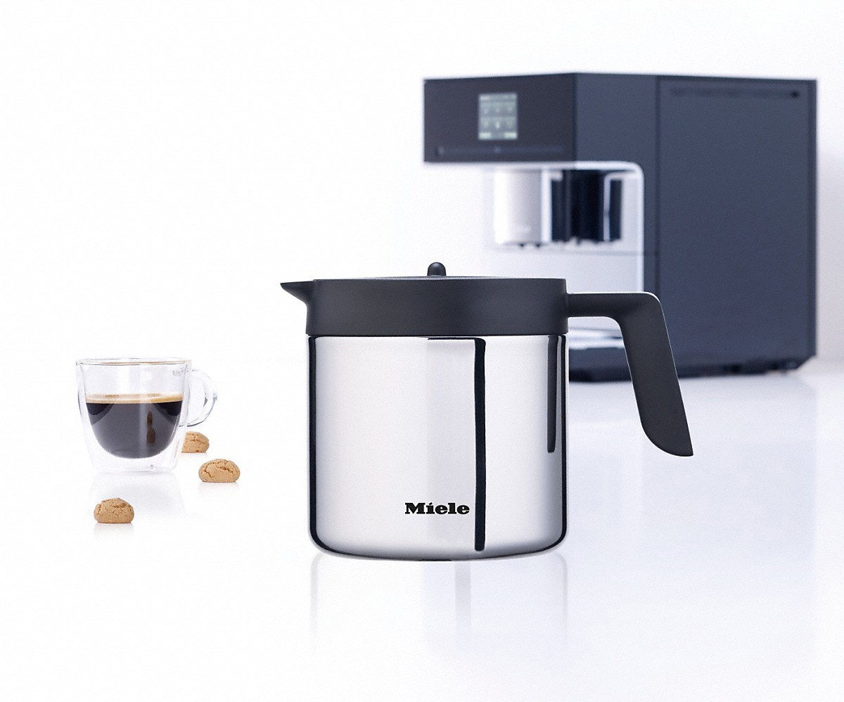 Eine Edelstahl-Kaffeekaraffe mit schwarzem Griff, neben einer Kaffeemaschine und einer Kaffeetasse.