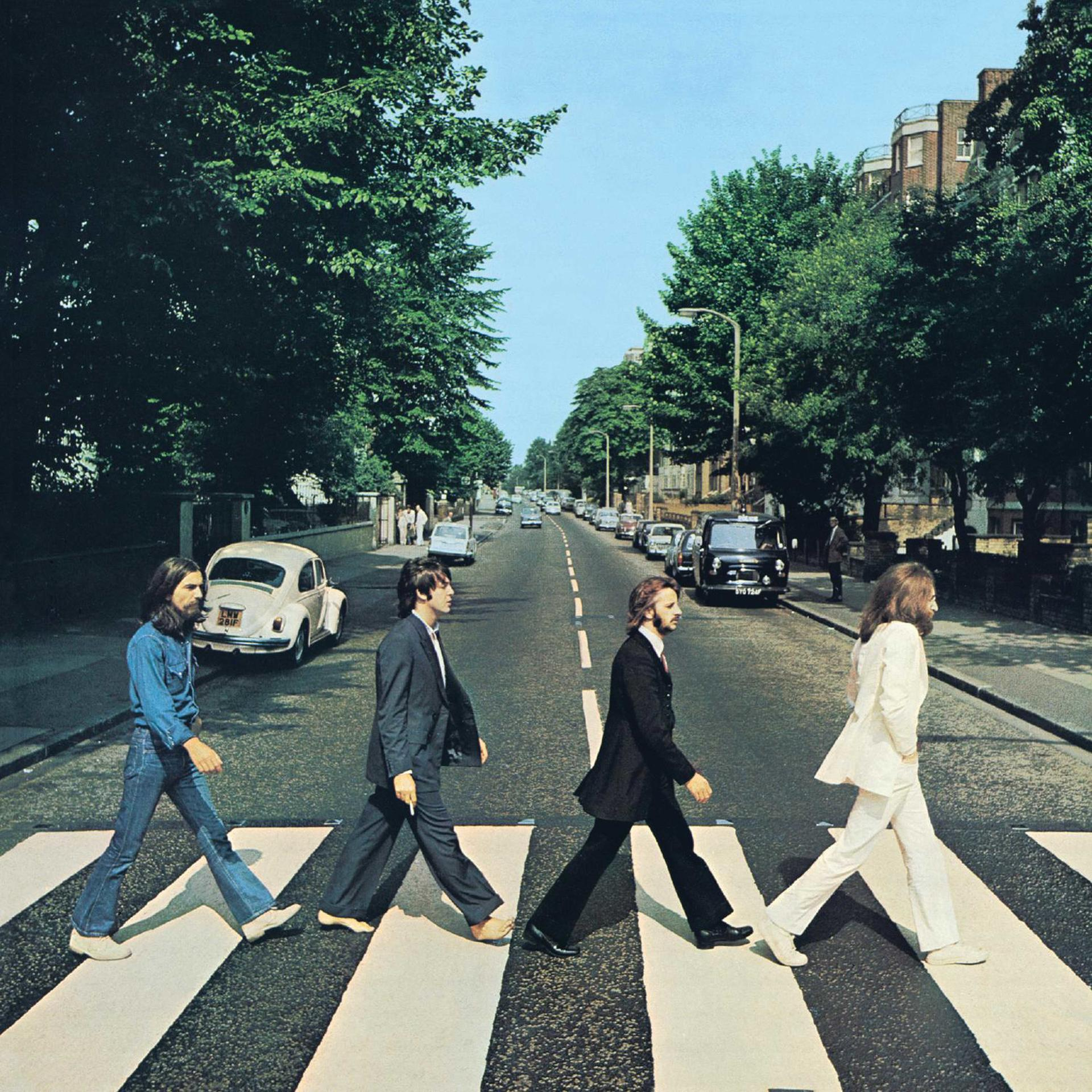 A Beatles az Abbey Roadon átkelve egy zebrán. Zöld fák szegélyezik az utcát, autók és épületek a háttérben.