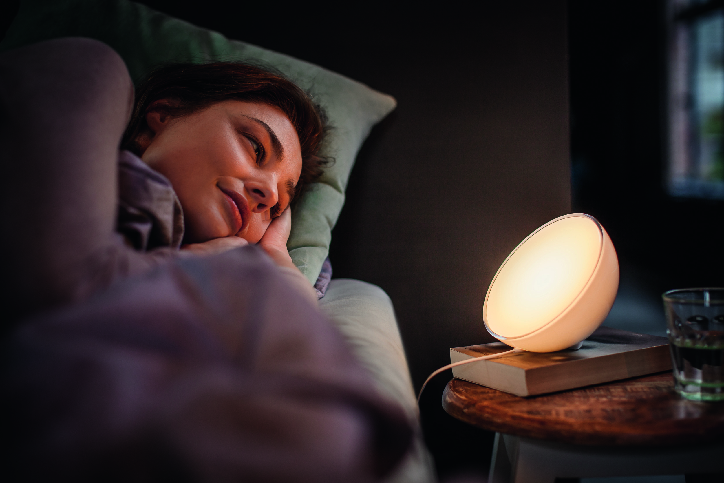 Eine Frau im Bett blickt auf eine weiße Nachttischlampe.