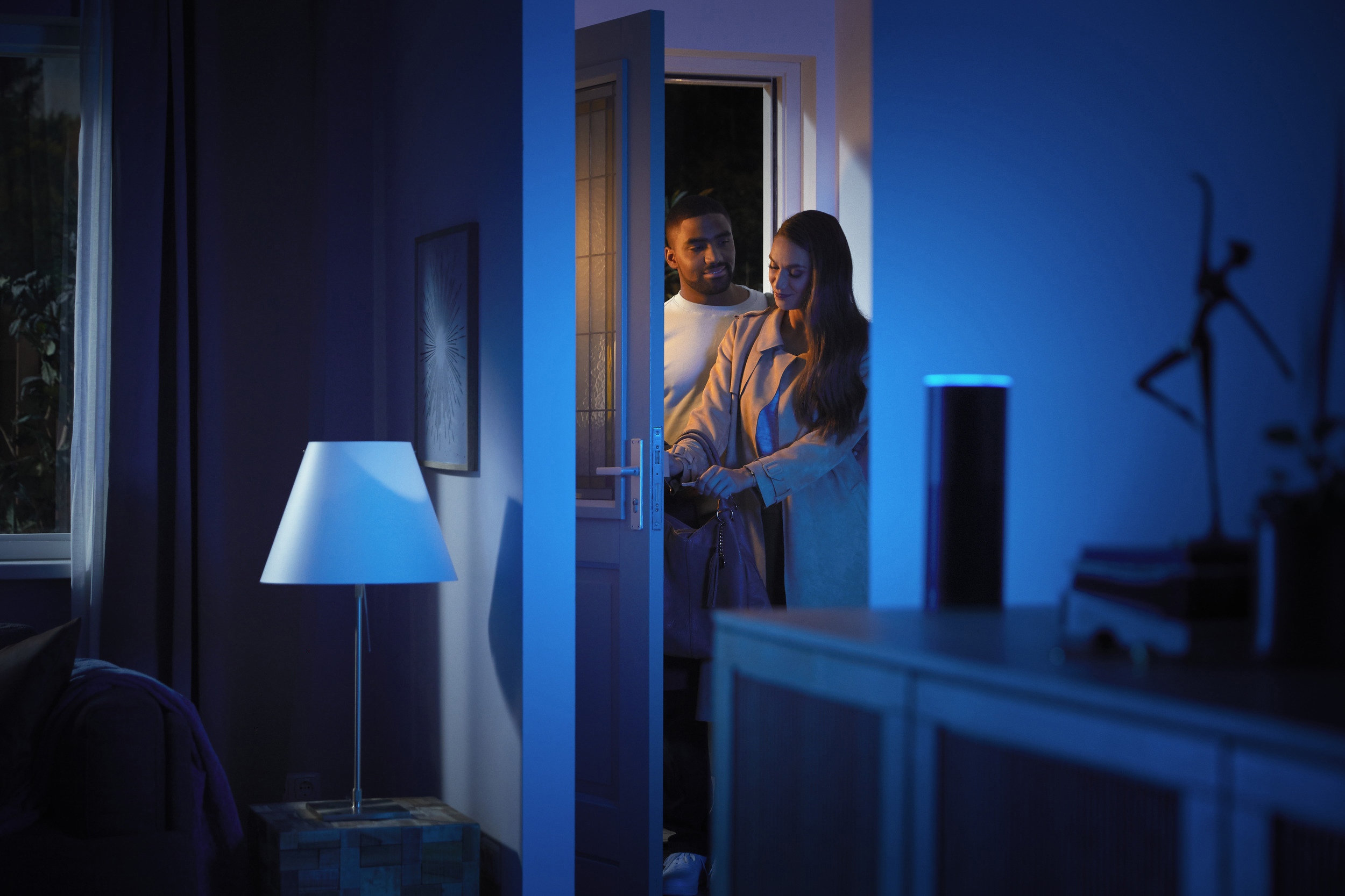 Ein Paar öffnet eine Haustür. Eine Lampe und ein Lautsprecher sind im Raum.