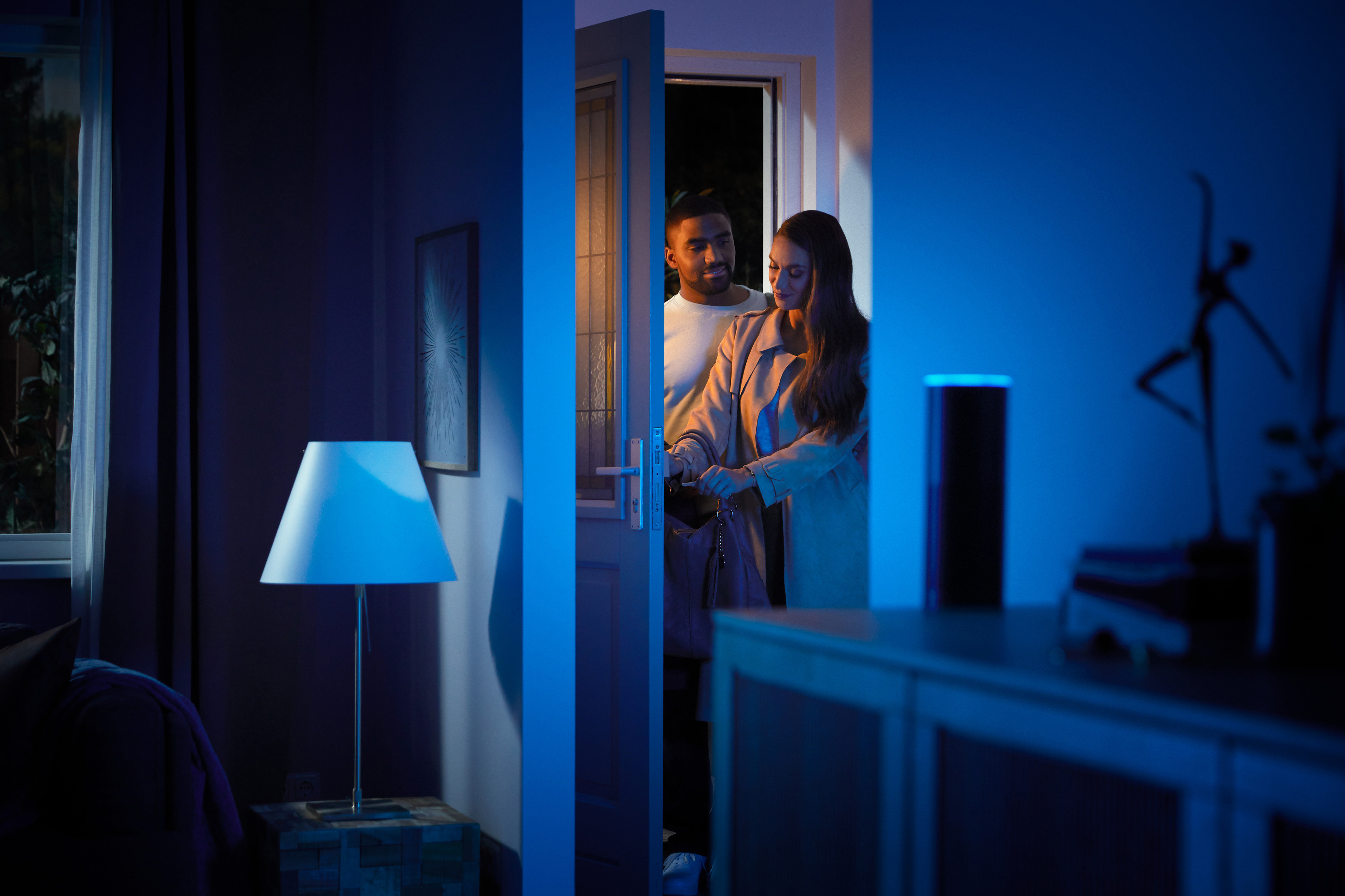 Paar betritt nachts ein Haus. Tür offen, Licht von innen, Smart-Home-Lautsprecher auf dem Beistelltisch.
