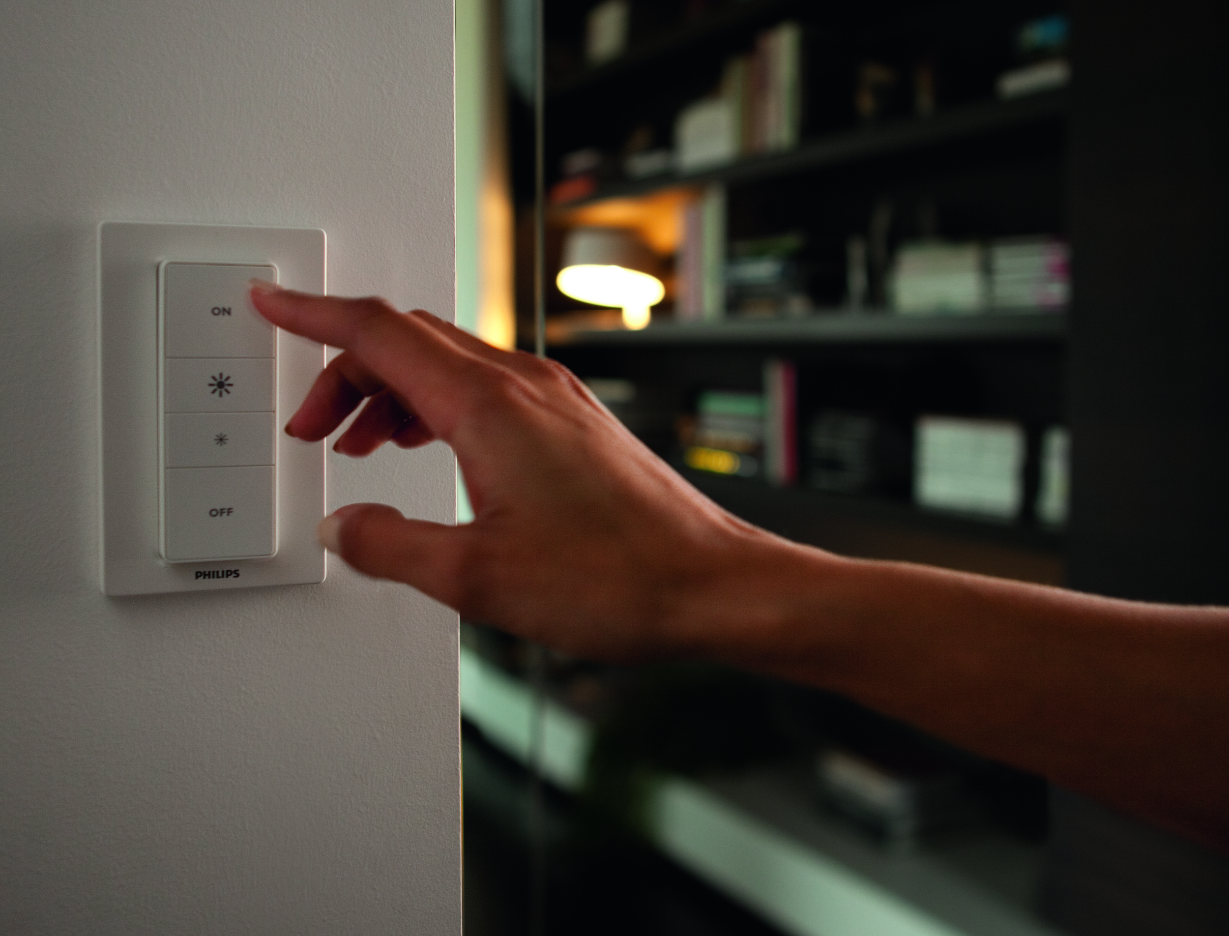 Eine Hand drückt die 'ON'-Taste an einem weißen, an der Wand befestigten Lichtschalter. Im Hintergrund ist ein Raum zu sehen.