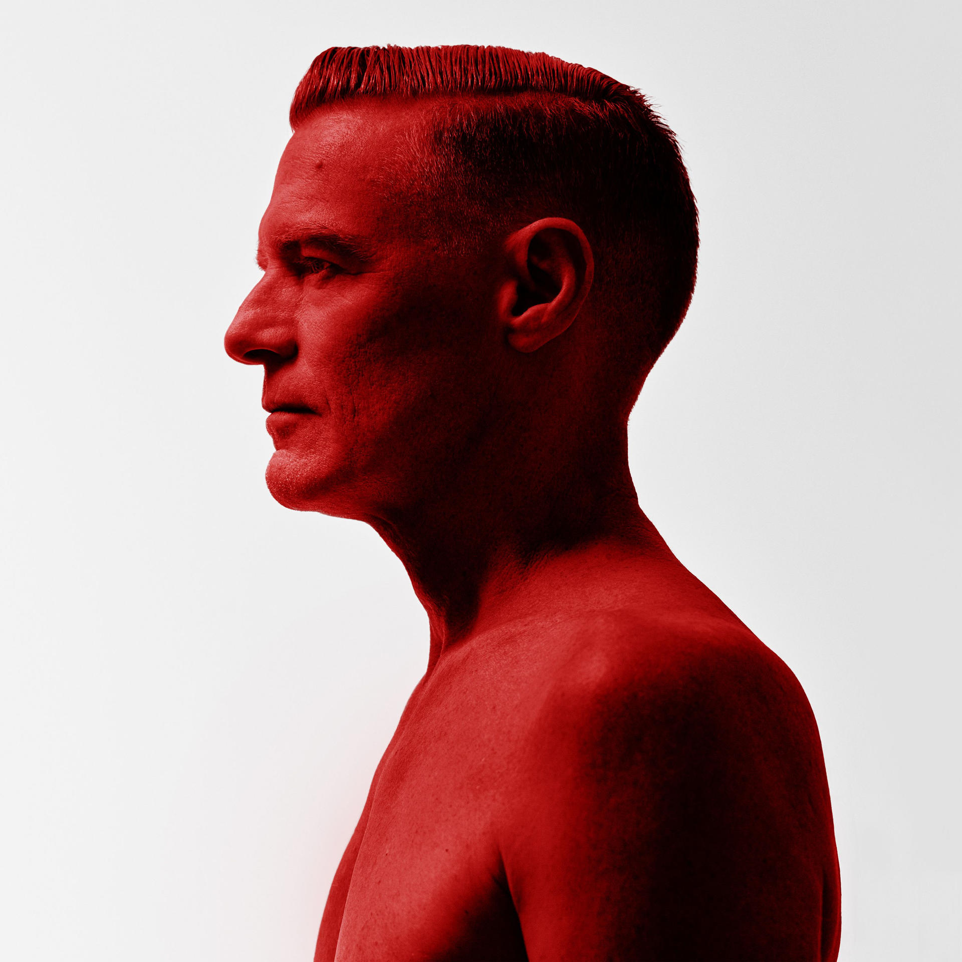 Ein Mann im Profil, rot getönt, mit rasiertem Kopf und nackten Schultern, vor weißem Hintergrund.