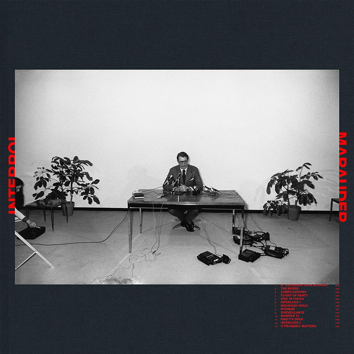 Albumcover: Interpol. Ein Mann an einem Tisch mit Mikrofonen, in einem schwarz-weißen Raum mit Pflanzen.