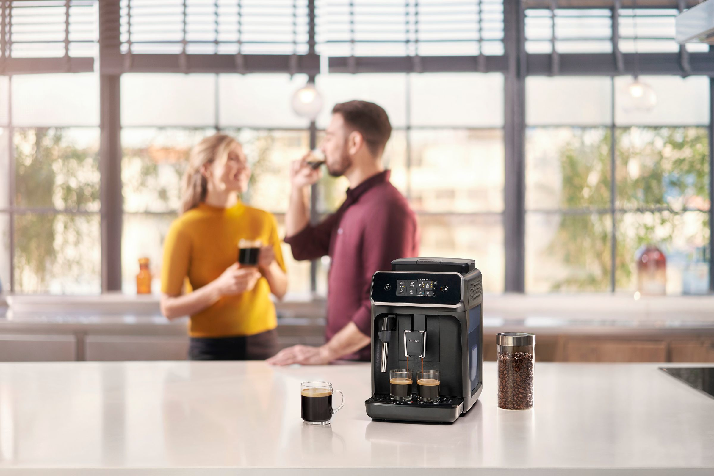 Ein Paar in einer Küche, mit Kaffeemaschine und Bohnen. Einer trinkt Kaffee.