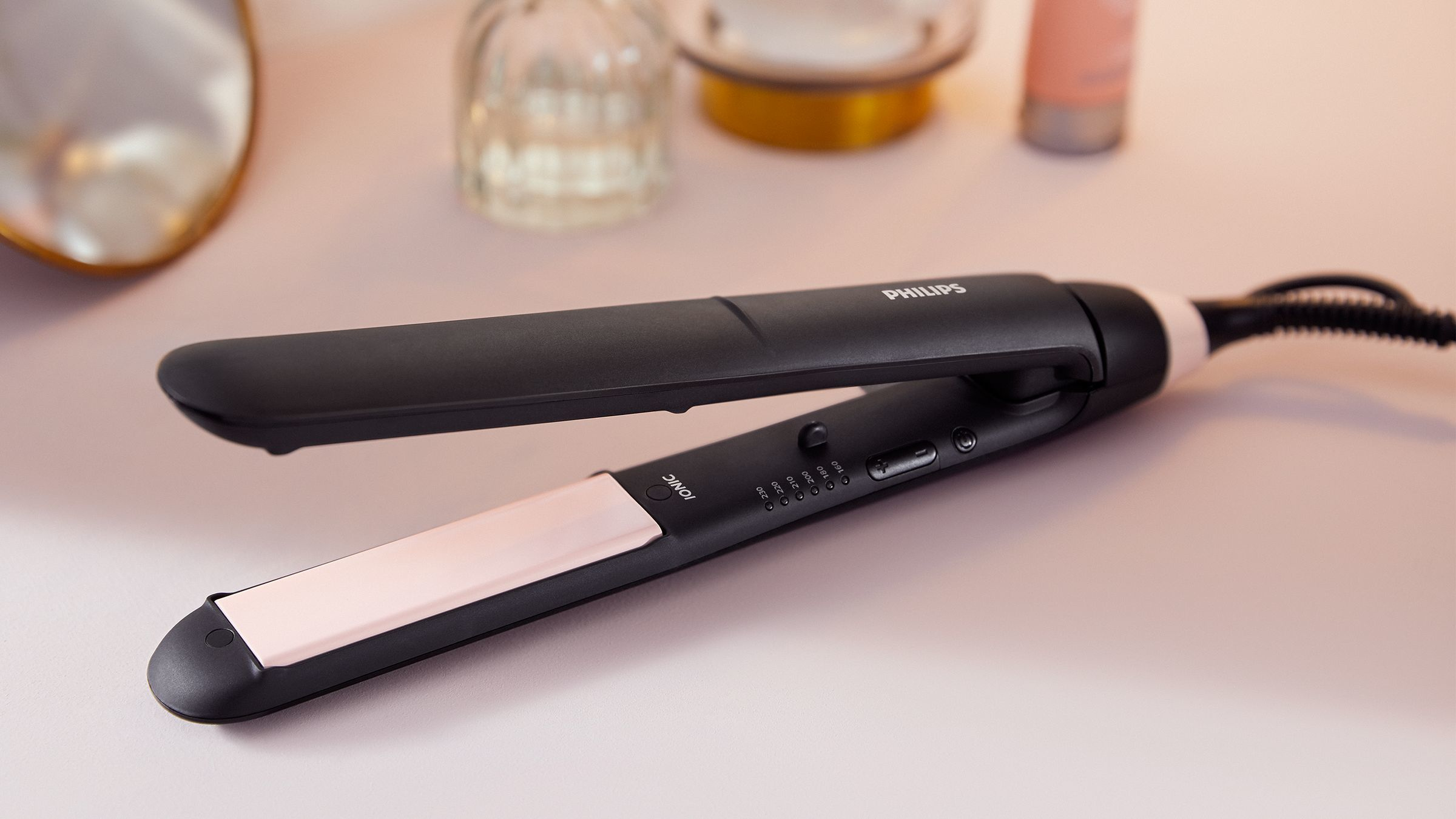 Ein schwarzer und roségoldener Philips Haarglätter auf einer rosa Oberfläche mit Schönheitsprodukten.