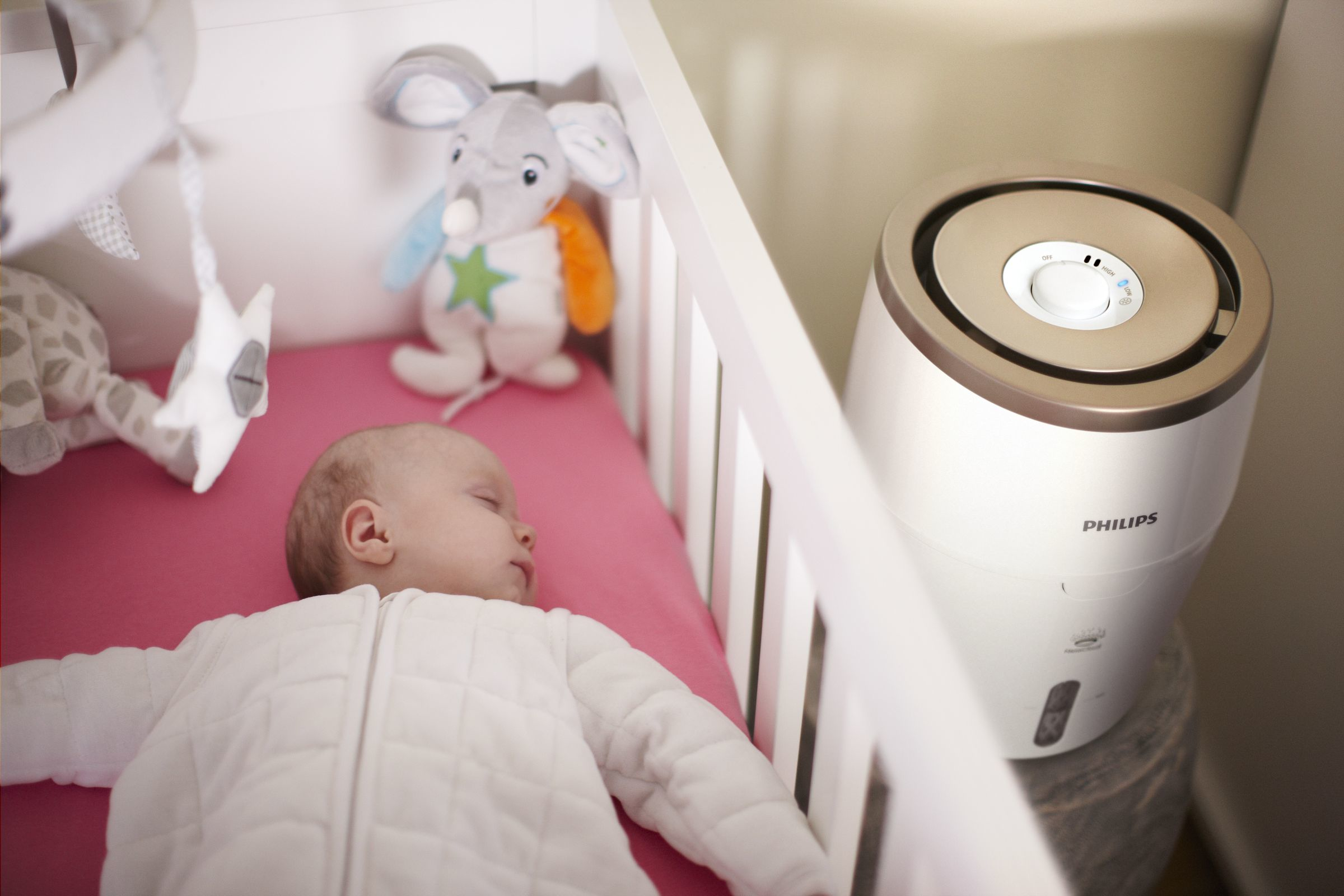 Ein Baby schläft in einem weißen Kinderbett, neben einem Luftbefeuchter und Spielzeug.