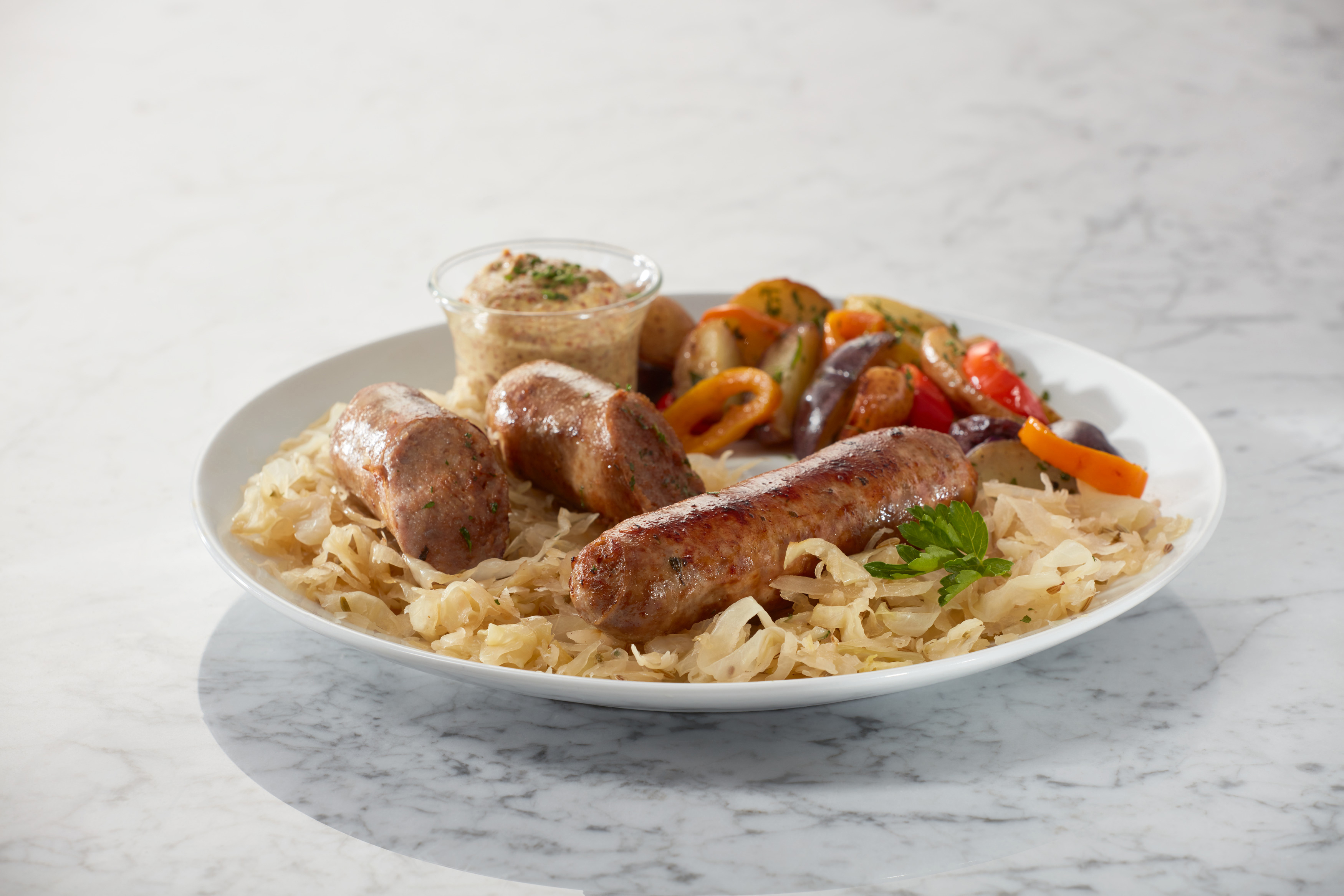 Teller mit Wurst, Sauerkraut, gebratenem Gemüse und Senf im Glas. Weiße Oberfläche.
