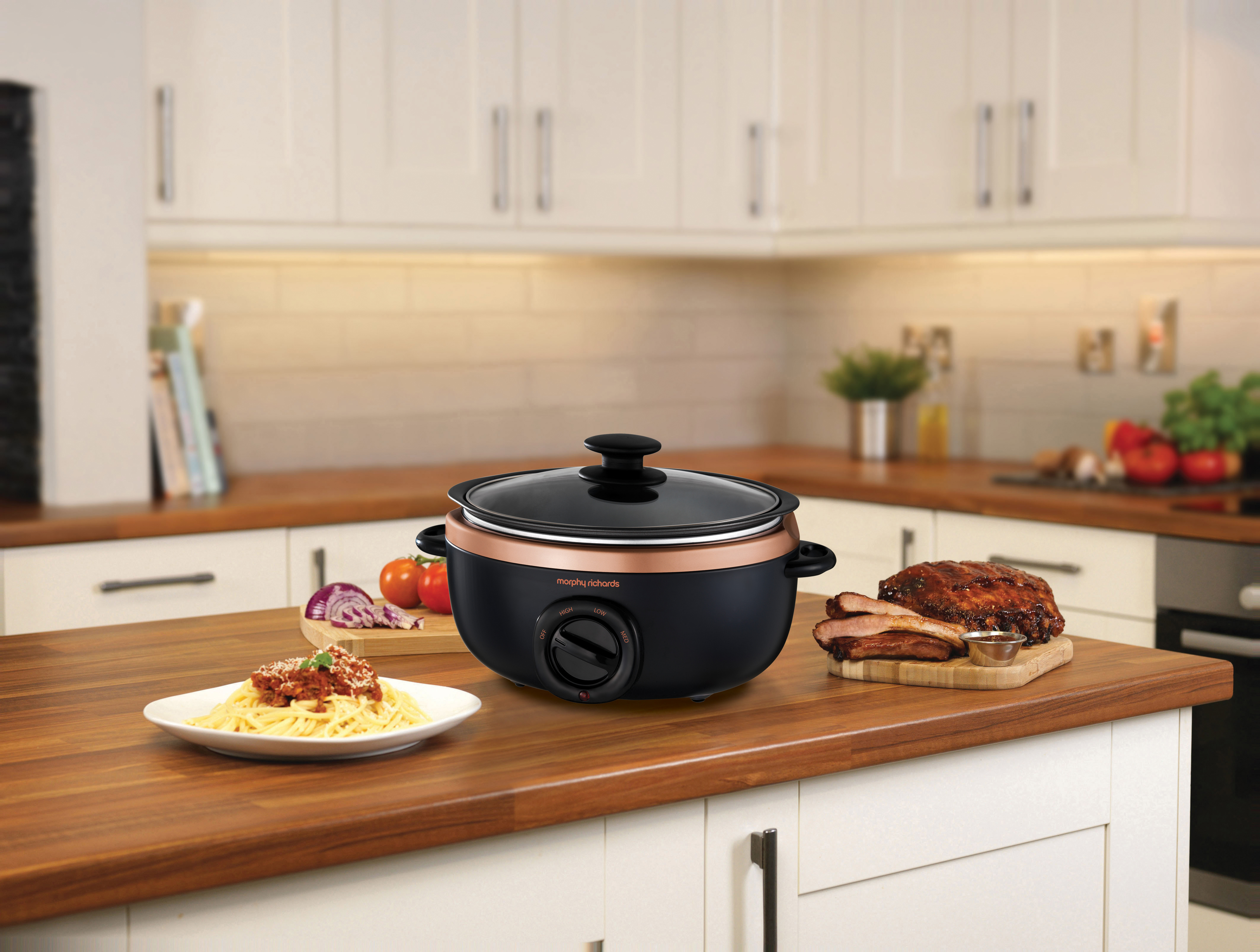 Ein schwarzer Slow Cooker mit Spaghetti, ein Schneidebrett mit Fleisch auf einer Holztheke.