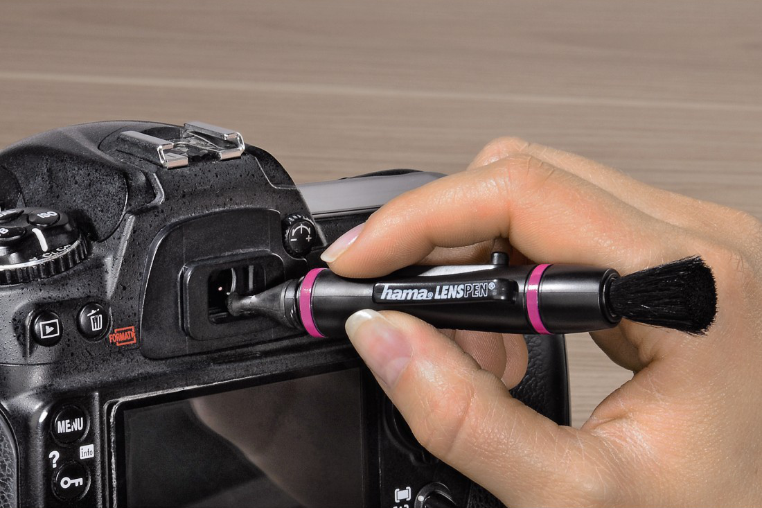 Une main nettoyant l'objectif d'un appareil photo avec un Lenspen. L'appareil photo est noir et argenté.