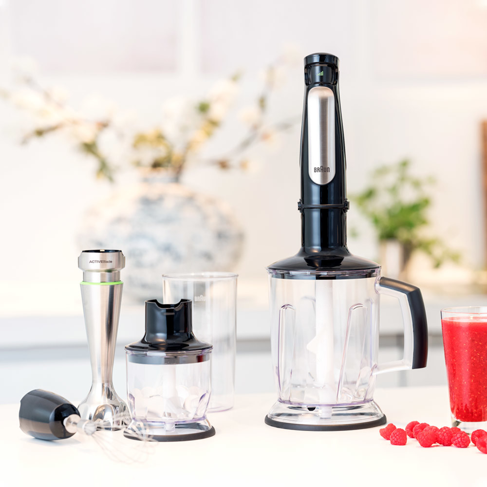 Ein Mixer mit mehreren Aufsätzen und einem Smoothie, mit einer Blumenvase im Hintergrund.