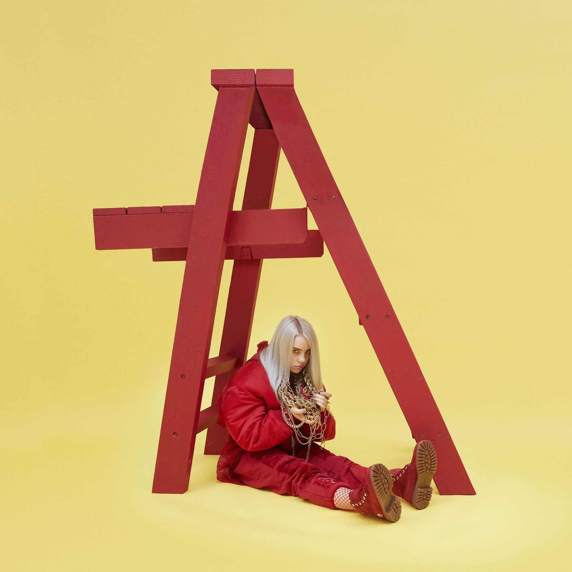 Billie Eilish est assise sous une structure en bois rouge, tenant des chaînes dorées. Fond jaune.