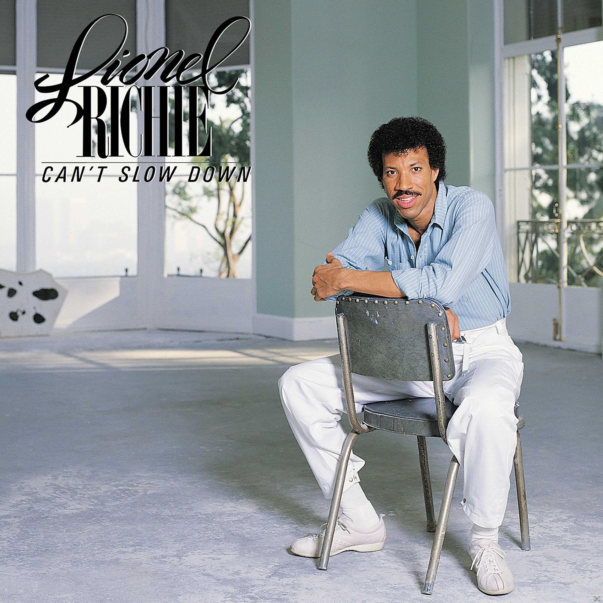Lionel Richie hátrafelé ül egy fém székek, kék inget és fehér nadrágot visel egy világos szobában.