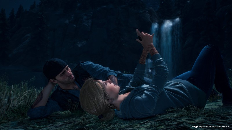 Una pareja acostada en el suelo, cerca de una cascada, mirando la pantalla de un teléfono.