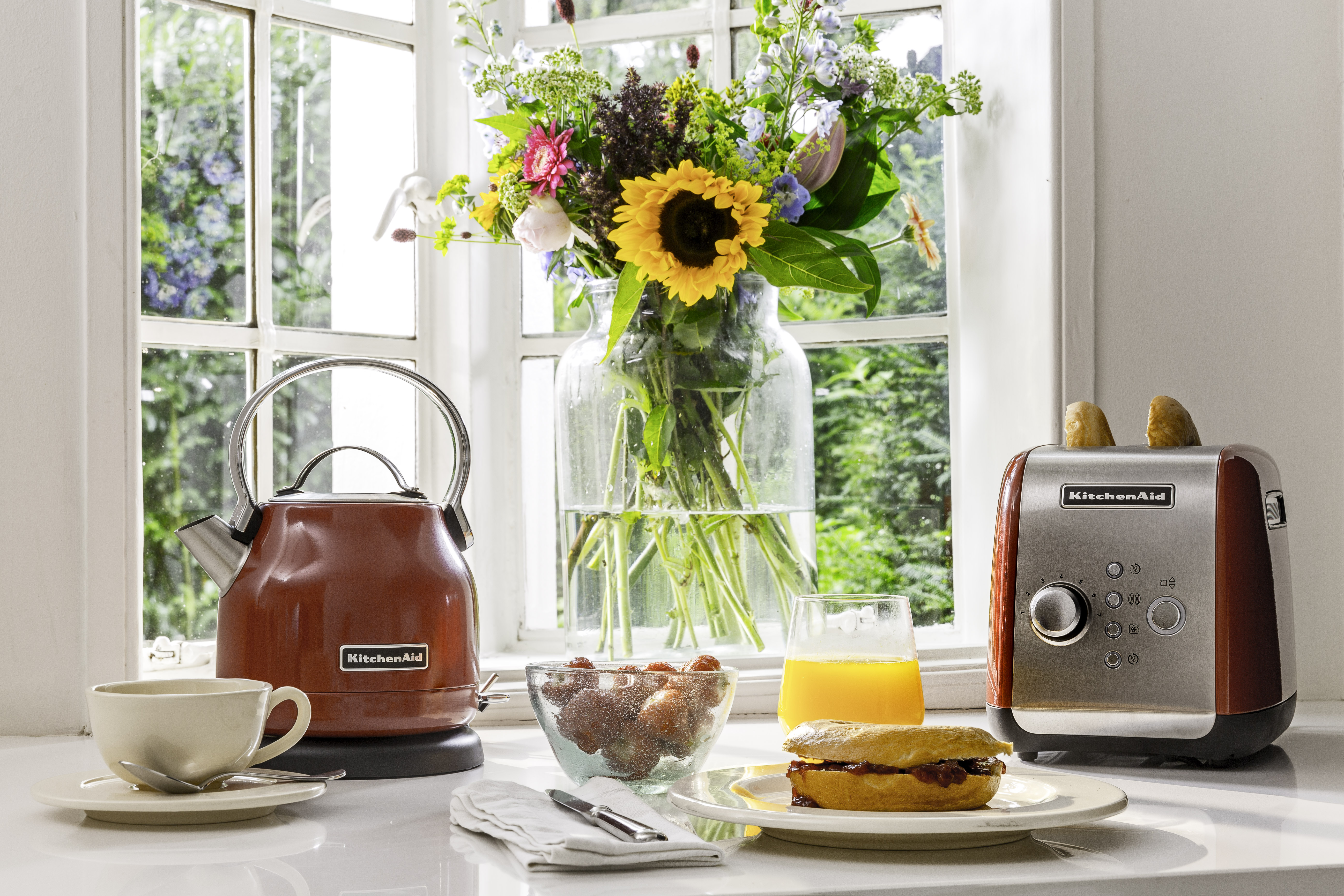 Eine Frühstücksszene am Fenster. Enthält einen Wasserkocher, Toaster, Blumen und Essen.