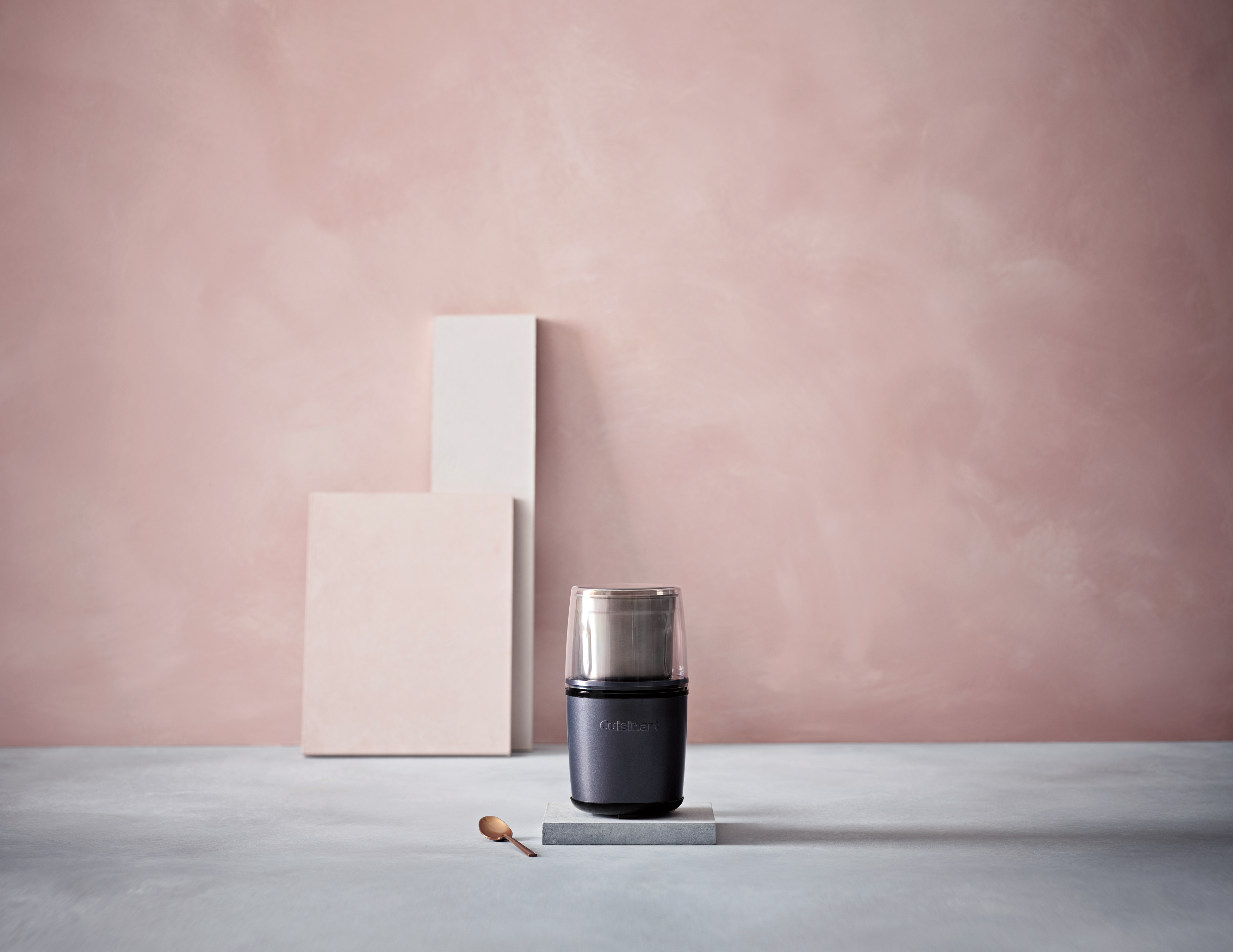 Dunkelgraue Kaffeemühle auf grauer Oberfläche, rosa Hintergrund, Holzlöffel.