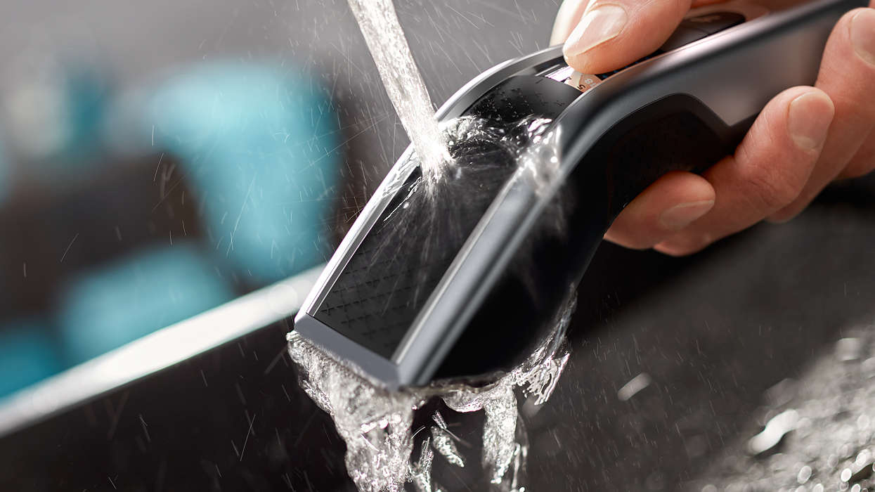 A black hair clipper being washed under running water. The water is flowing over the blades of the device.