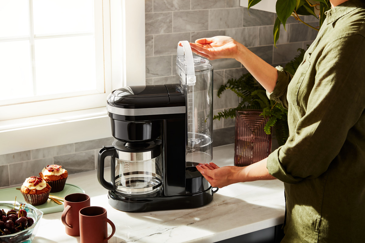 Eine Frau, die Wasser in eine schwarze Kaffeemaschine mit Glaskaraffe und Muffins füllt.