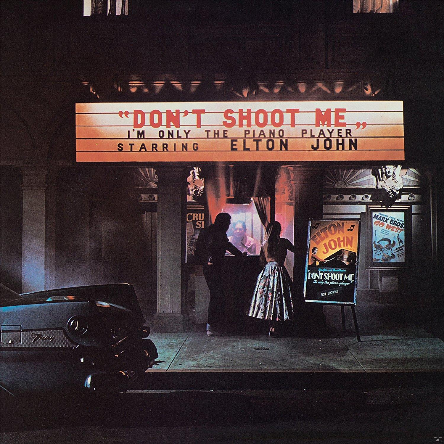 Egy színházi felirat a következőt írja: "Don't Shoot Me", Elton John főszereplésével. Emberek egy plakát közelében, balra egy autó.