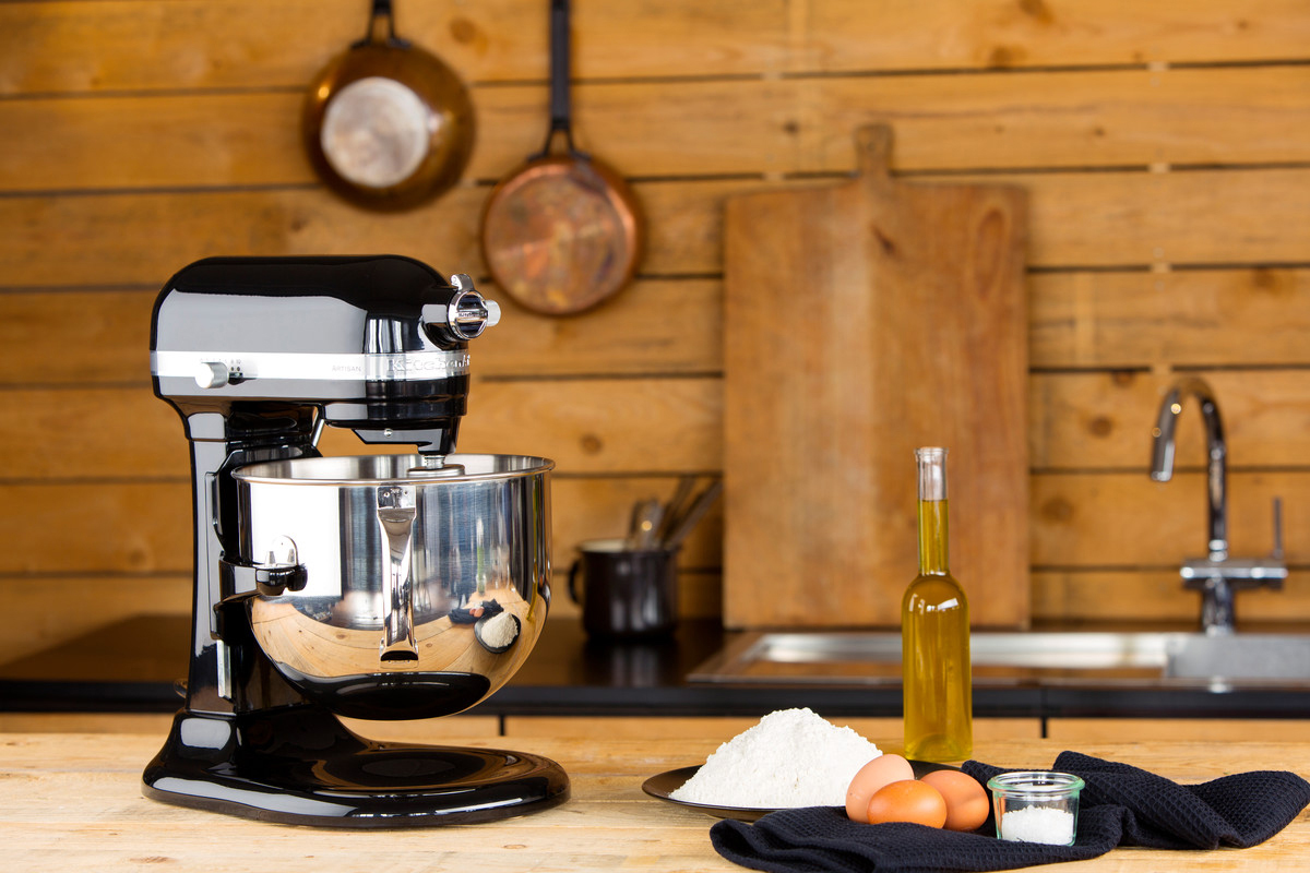Schwarzer KitchenAid-Mixer mit Schüssel, Mehl, Eiern, Öl auf einem Holztisch. Holziger Hintergrund.